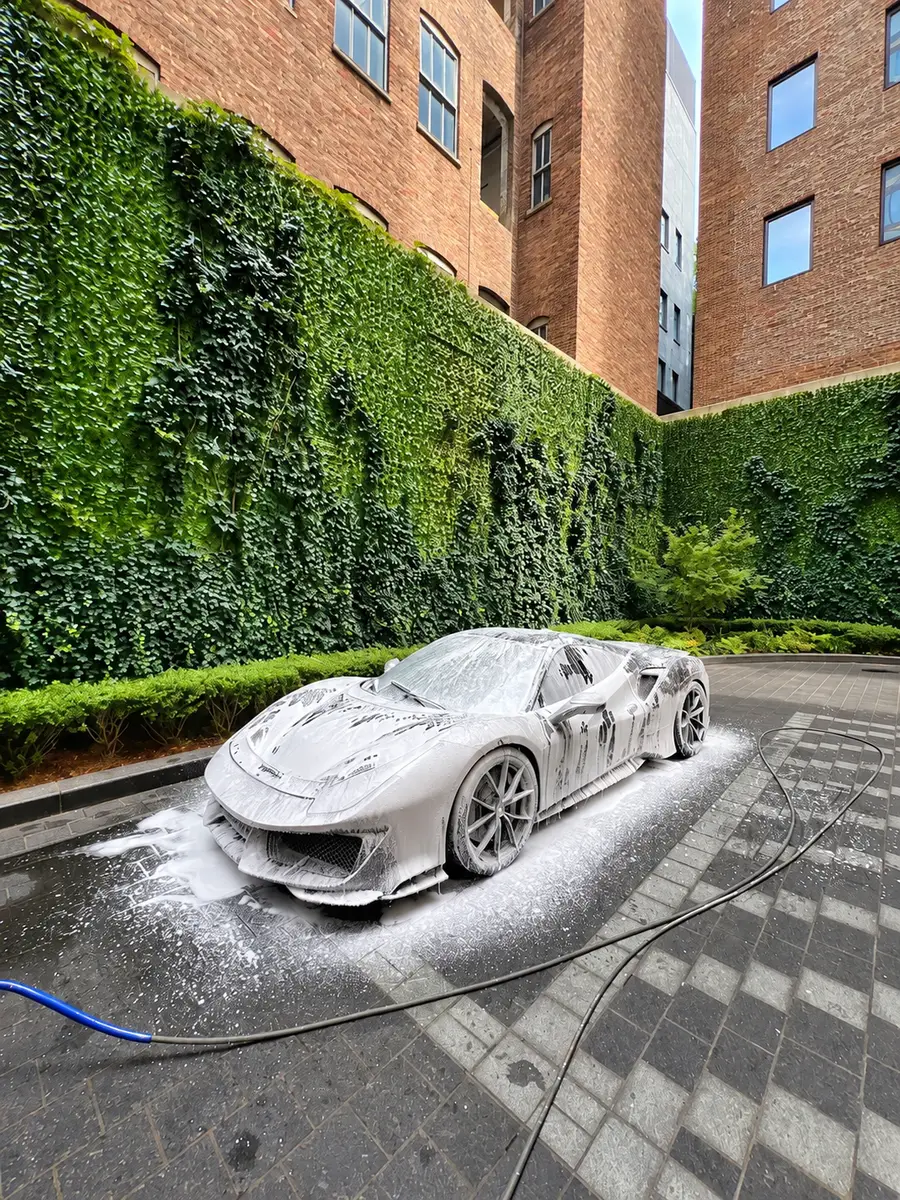 White sports car covered in foam during a hand wash in a brick courtyard, pressure washer hose on the pavers
